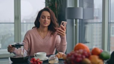 Kahvaltıda akıllı telefon kullanan bir kadın rahat ev mutfağında fincana kahve dolduruyor. Rahat neşeli kız masada cep telefonuyla mesaj okuyor. Mutlu bayan öğle yemeğini evde yiyor.