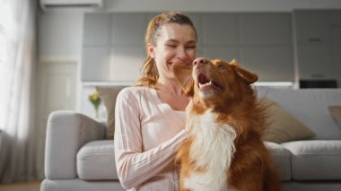 Evde gülümseyen sevimli bir köpeğe sarılan bir kadın. Modern dairede tüylü evcil hayvanı okşayan neşeli, güzel bir kız. Dişi köpek sahibi içeride sevimli hayvanlara sarılırken eğleniyor..