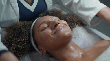 Gülümseyen kadın güzellik prosedüründen sonra masajdan zevk alıyor. Eller nemlendirici kremi pürüzsüz cilt üstü görüntüsüne sürer. Mutlu Afro-Amerikan Spa Sağlık Kliniği 'nde kimliği belirsiz kozmetik uzmanı konuşuyor..