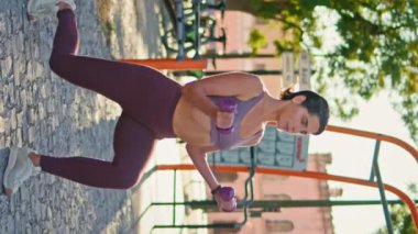 Kaslı sporcu kadın, sokak antremanında dambılları dikey kaldırarak çömeliyor. Odaklanmış olgun bir kadın tek başına açık hava spor salonunda ağırlık kaldırma eğitimi alarak çömelme hareketleri yapıyor. Sağlıklı yaşam tarzı kavramı 