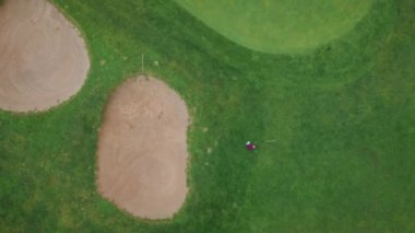 Hava manzaralı golfçü, sığınaklar tarafından çerçevelenmiş canlı yeşil sahada sallanıyor. Çimenler, doğayla çelişen çimlerle doludur. Yeşillik oyun alanını kaplıyor. Golf oyuncusu sakin bir oyun oynuyor.