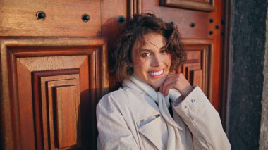 Neşeli kız rüzgarlı bir hafta sonu pozu veriyor ahşap kapıya yaslanıyor ve neşe verici bir yakınlaşma sergiliyor. Zarif montlu kıvırcık saçlı kadın dokulu kapının önünde dikiliyor. Kentsel alanda poz veren gülümseyen bir model.