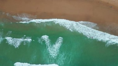 Sörfçüler, dinamik okyanus manzarasında dalgalar halinde yüzerler. Hava manzaralı turkuaz deniz suyu güzel kumlu sahili çok yavaş çekimde yıkıyor. Kıyı yaşam tarzı ve su sporları coşkusu