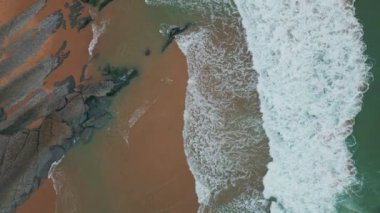 Köpüklü dalgalar, kayalık kıyı şeridi manzaralı kumlu sahili yıkıyor. Tortu ve erozyon desenleri turkuaz deniz suyuyla harmanlanmış. İnsansız hava aracı okyanus dalgalarının engebeli kıyılarla buluşmasını çok yavaş çekimde çekti.