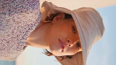 Sakin bir turist, deniz kıyısında dikey çekimde poz veriyor. Huzurlu güzel kız deniz kenarında tek başına dinleniyor. Güneş ışığında yalnız genç bir kadın kumsalda kameraya bakıyor. Gezgin gün batımının tadını çıkarıyor.