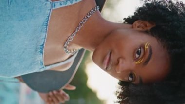 Kaykay tahtası tutan hippi kız. Arsız gülüşlü kameraya benziyor. Güzel Afro-Amerikalı genç kaykaycı parkında dikey video ile poz veriyor. Ayakta duran kıvırcık kaykaycı