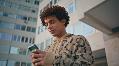 Modern şehrin arka planında duran akıllı telefonu kullanan odaklanmış bir adam. Kıvırcık saçlı genç, şehir ortamında sosyal medyada cep telefonuyla geziniyor. Latin hipster şehir caddesinde cep telefonuyla mesajlaşıyor..