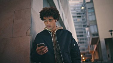 Zoomer cep telefonu, şehir gece atmosferi tarafından aydınlatılıyor. Kapüşonlu İspanyol adam şehir geçmişinde akıllı telefon mesajları yolluyor. Kulaklıklı kıvırcık erkek, dışarıda cep telefonu ekranına bakıyor..