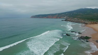 Okyanus dalgaları kıyı bulutlu hava manzarasına çarpıyor. Çok yavaş çekimde turkuaz suya sallanan sörfçülerin tepesindeki engebeli uçurum kulesi. Mavi deniz dalgası güzel kumsalda süzülüyor beyaz köpük yapıyor.