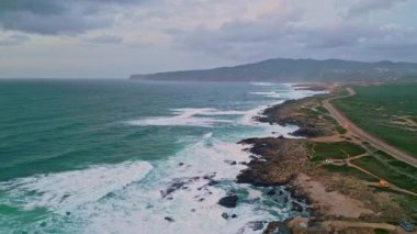 Hava manzaralı engebeli kıyı şeridi çarpışan turkuaz dalgalarla. Vahşi yeşil arazi sınırı güzel fırtınalı okyanus manzaralı manzaralar yaratıyor. Asfalt yol kıyı şeridinde uzanıyor. Dramatik sahil güzelliği.