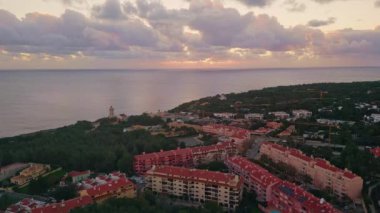 Sahil kasabasının üzerindeki alacakaranlık gökyüzü sakin ve sakin okyanus yüzeyiyle çevrili. Deniz feneri yemyeşil ile kaplanmış sıcacık deniz kenarı şehrine inen hava manzaralı bir akşam. Bulutlu gökyüzü deniz manzarası üzerinde