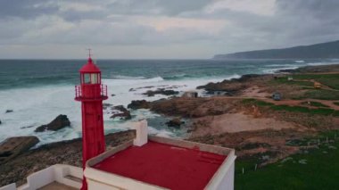 Deniz feneri, bulutlu akşam hava manzarasına fırtınalı deniz manzarası yerleştirdi. Dalgalar dramatik gökyüzünün altındaki kayalık kayalıklara çarpıyor. Çalkantılı ve köpüklü deniz suyuna bakan kırmızı sahil feneri güvenli seyrüsefer sağlıyor..