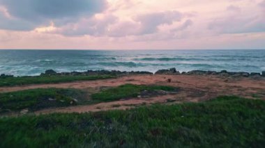 Köpüklü dalgalar, kasvetli gökyüzü drone görüşü altında kıyı şeridini çökertiyor. Fırtınalı turkuaz dalgalar tarafından süper yavaş çekimde yıkanan güzel çimenli arazi kıyısı. Okyanus suyu engebeli sahil doğasına sıçrıyor..