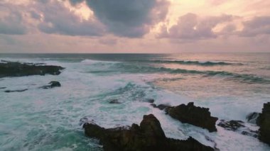 Panoramik manzara okyanus dalgaları bulutlu gündoğumu gökyüzü altında. Altın sabah ışıltısı yaban deniz manzarası yumuşak ışık drone çekiminde. Fırtınalı deniz suyu köpükle sıçrıyor kıyı kayalarını süper yavaş çekimde eziyor..