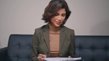 Deri koltukta kağıtları analiz eden profesyonel bir kadın. Şık ofis ortamında belgeleri dikkatle okuyan odaklanmış bir iş kadını. Modern klinik ziyaretçisi panodaki bilgilere bakıyor.