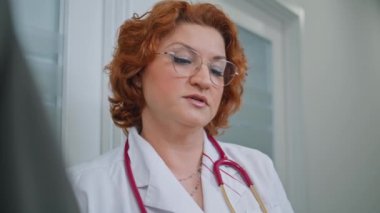 Klinikteki sağlık sigortası prosedürü anlaşmasını açıklayan tıbbi danışman. Gözlüklü kızıl saçlı kadın ofiste bilinmeyen bir hastayla konuşuyor. Paltolu profesyonel, sağlık hizmeti tavsiyesi veriyor..
