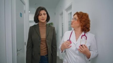 Hastane koridorunda hastayla sağlık hakkında konuşan bir uzman var. Beyaz önlüklü doktor klinikte yürüyen kadına danışmanlık yapıyor. Kızıl saçlı doktor ofis koridorunda tıbbi danışmanlık yapıyor..