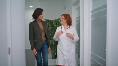 Klinik koridorunda hastayla birlikte yürüyen bir tıp uzmanı. Kendine güvenen doktor modern koridorda sağlık hizmetlerini tartışıyor. Gülümseyen esmer kadın sağlık sigortası uzmanından sağlık danışmanlığı istiyor.