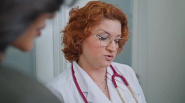 Kendine güvenen hekim, modern ofis kapanışında duran klinik ziyaretçisine sağlık danışmanlığı tavsiyeleri veriyor. Profesyonel kadın doktor koridorda kadın hastaya danışmanlık yapıyor. Tedaviyi açıklayan sağlık danışmanı