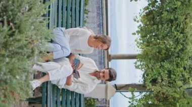 Park bankının dikey videosunda gülen çiftin kitap okuması. Romantik şehir buluşmasında esmer adam kız arkadaşını kucaklıyor. Mutlu çift yeşillik meydanında eğleniyor. Aşk konsepti