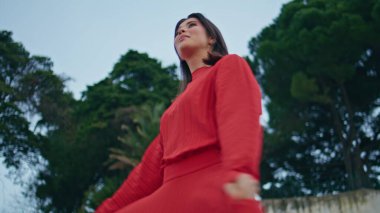 Mutlu esmer kadın parkı tek başına geziyor. Kırmızı elbiseli pozitif kadın sıcak şehir bahçesinde kamera kıvrımlı görünüyor. Kaygısız bayan akşam dinleniyor. Neşeli bir kadın.