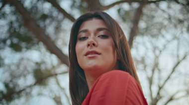 Portre esmer kız tek başına gülümsüyor. Mutlu, neşeli bir kadın yaz güneşinin altında gülümseyen bir kamera görüyor. Kırmızı elbiseli güzel bayan yeşil parkta yalnız poz veriyor. 