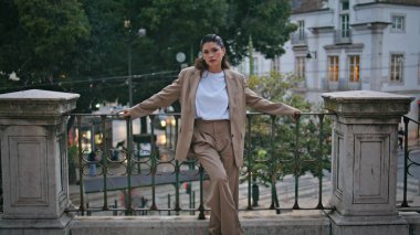 Metal korkuluklara dayanan şık bir model. Güzel şehirde poz veren rahat bir kadın şık bir takım elbise giyiyor. Çekici kız sakin bir şehirde hafta sonları dinleniyor..