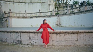 Şık model dinlenme çeşmesi sınırı eski kasabada duruyor. Tek başına poz veren kırmızı elbiseli muhteşem düşünceli kız. Genç esmer bayan sakince şehir parkında yürüyor. Halk yaşam tarzı konsepti