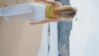 Arka manzaralı kumsalda yürüyen kadın güzel bir akşamın tadını çıkarıyor. Tanımlanamayan genç bir kız boş yaz deniz kıyısında tek başına dikey videoda geziniyor. Beyaz pantolon giyen kıvırcık sarışın, kıyıda dinleniyor.