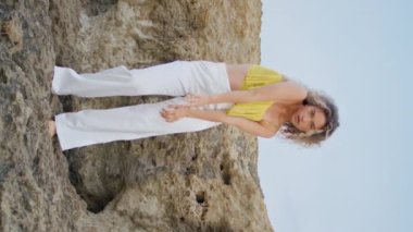 Kıvırcık kız, akşam dikey görüntüsünde kumlu kayanın üzerinde dans ediyor. Esnek, genç bir kadın. Vücudunu hareket ettiriyor. Doğanın tadını çıkarıyor. Profesyonel dansçı, taşlı arazide çağdaş bir tarz sergiliyor..