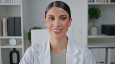 Poliklinikte gülümseyen kameralı dost canlısı terapist portresi. Modern klinikte oturan beyaz cüppeli neşeli kadın doktor. Sağlık merkezinde poz veren bir tıp çalışanı.