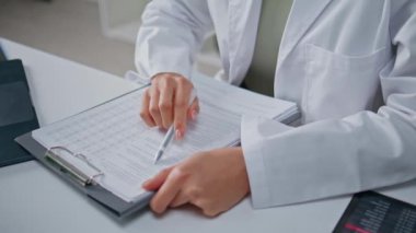 Sağlık uzmanı klinik masasındaki tıbbi belgelerde bilgileri kontrol ediyor. Bilinmeyen kadın doktor hastane işyerinde sigorta formu dolduruyor. Beyaz cüppeli doktor rapor yazıyor..