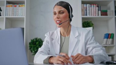 Kulaklık doktoru laptoptaki kamerayla konuşuyor. Klinikteki hastalarla iletişim kuruyor. Çevrimiçi doktor uzaktan tıbbi destek sağlıyor. Kulaklıklarda sağlık danışmanı. Belgedeki verileri kontrol ediyor.