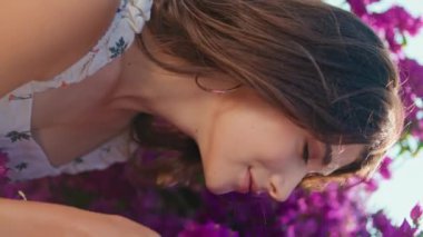 Romantik kız, rüzgârlı yaz sabahı dikey çekimde menekşe çiçeklerini kokluyor. Sıcak bir günde gülümseyen çiçeklerin tadını çıkaran mutlu esmer kadın. Çekici, neşeli kadın bahçede siesta yapıyor.