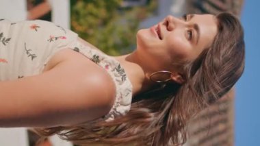 Güneş ışığı altında dikey çekimde eski şehirde gezinen tatmin olmuş esmer kadın. Gülümseyen kadın saçlarına dokunuyor sıcak yazın tadını çıkarıyor. Çekici, rahat, romantik elbiseli kız. Manzara seyrediyor. Kadın yürüyüşü