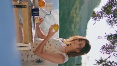 Dağ manzaralı terasta meyve hazırlayan sakin bir model. Gülümseyen mutlu kadın üst balkonda yemek yaparken elma kesiyor. Neşeli kız yeşillik köyünde tek başına yemek yapıyor.