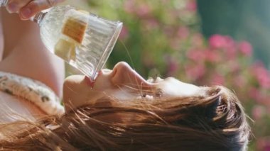 Limonata yudumlayan sakin kız yaz terasında dikey çekimde dinleniyor. Rüzgarlı saç stili kadın sağlıklı su içeceği içiyor ve kahvaltısını temiz havada yapıyor. Duygusal model haftasonu doğada