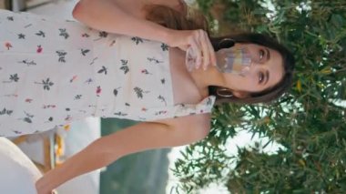 Dinlenme modeli limonata tutuyor. Doğa terasında dikey çekimde içki içiyor. Romantik bayan mesafeli görünüyor. Sadece meyvelerle su içiyor. Nazik kadın soğuk sıvı yudumluyor balkonda rüzgarı hissediyor