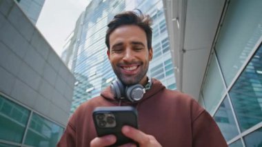 Ofis bölgesinde gülümseyen adam akıllı telefondan mesaj atıyor. Yakın plan neşeli adam modern şehir manzarasında cep telefonu kullanıyor. Kulaklıklı mutlu teknoloji erkeği açık havada sohbet etmekten hoşlanıyor..