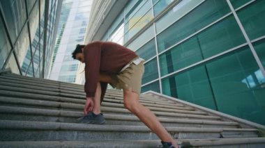 Şehir koşucusu spor ayakkabılarını modern cam mimarisi düşük açıyla bağlıyor. Atletik adam dışarıda spor yaparken ayakkabılarını düzeltiyor. Enerjik sporcu adam sabah koşa koşa yukarı çıkıyor..