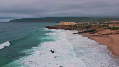 Huzurlu sahil alacakaranlıklarında köpüklü dalgalara bakan Cliffside Otel. İnsansız hava aracı akşam panorama kıyısı köpüklü deniz tarafından ağır çekimde yıkandı. Rocky kıyı şeridi manzaralı seyahat rotasında okyanusu çerçeveliyor.
