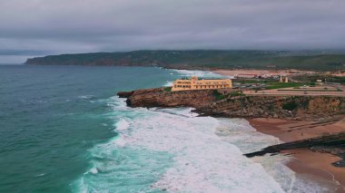 Alacakaranlık İHA 'sının vurduğu kayalık plajda duran sahil binası. Engebeli kayalıklara çarpan turkuaz okyanus dalgalarıyla manzaralı sakin deniz manzarası. Akşamları dalgalar tatil kaçamağında kayalık kıyıları yıkar..