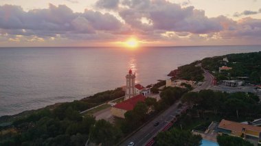 Okyanusa bakan deniz feneri güneş doğumu drone manzaralı. Sakin okyanus yüzeyine yansıyan portakal rengi güneş ışığı. Sahil şeridine kurulmuş kıyı kasabası yemyeşil yemyeşil. Deniz ufkundan yükselen güneş.