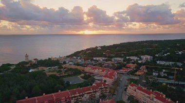 Sahil kasabası, deniz fenerinin yanında gün batımında parlıyordu. Hava manzaralı sakin deniz manzaralı yerleşim yerleriyle buluşuyor. Huzurlu akşam ışığı manzarayı güzelleştiriyor. Kıyı şeridindeki konforlu çatı binaları