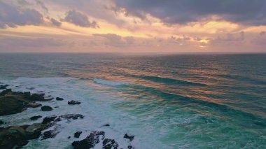 Okyanus yüzeyi altın akşam ışığı drone görüntüsünü yansıtıyor. Deniz dalgaları kıyı uçurumlarına çarpıyor. Nefes kesen günbatımı gökyüzü altında. Manzaralı turkuaz sörf suyu süper yavaş çekimde taştan kıyı şeridine yuvarlanıyor..