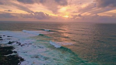 Deniz gündoğumu okyanus dalgaları üzerinde kıyı uçurumlarının hava manzarasına çarpıyor. Ufuk, altın ışıkla parıldıyor. Huzurlu deniz manzarası. Köpüklü deniz sörfü taşların üzerine çok yavaş bir şekilde vuruyor..