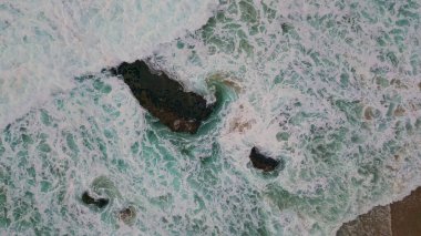 Okyanus dalgaları kıyı manzarasında fokurduyor. Kumlu sahilde su çok yavaş çekimde köpürüyor. Fırtınalı deniz sörfü kayalık kıyı şeridini yıkıyor beyaz köpük yapıyor. Resimli deniz manzarası.