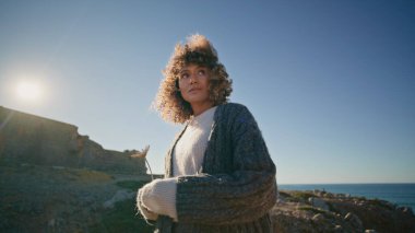 Romantik model, sonbahar rüzgârlı havalarda deniz manzarasını yakınlaştırıyor. Çiçeği tutan zarif çekici kadın taşların arasındaki mesafeyi arıyor.