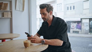 Rahat bir adam kafeteryada oturmuş akıllı telefon ekranına yakından bakıyor. Genç adam kafeteryada sosyal medya izlemek için cep telefonu kullanıyor. Kahve dükkanında sakallı yakışıklı bir erkek..