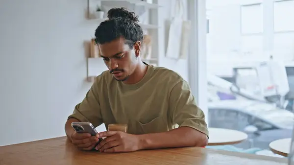 stock image Latin man sitting cafe table looking smartphone screen alone close up. Curly brazilian hipster scrolling mobile phone browsing social media. Male freelancer messaging at cozy coffee house interior. 